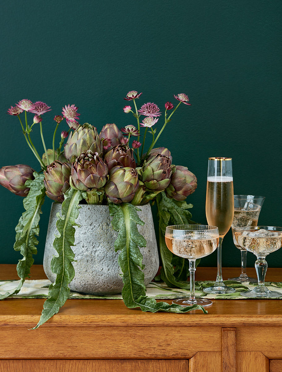 Coupes et flûte de champagne rosé près d'un bouquet de fleurs d'astrances et artichauts, fond vert forêt