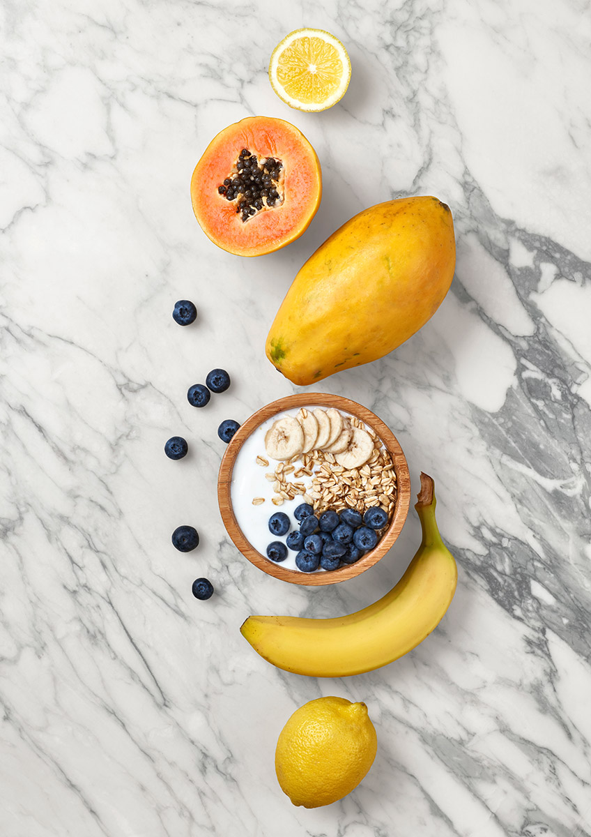 Flat Lay bol muesli et fruits detox : citron, banane, myrtille, papaye, sur un fond de marbre